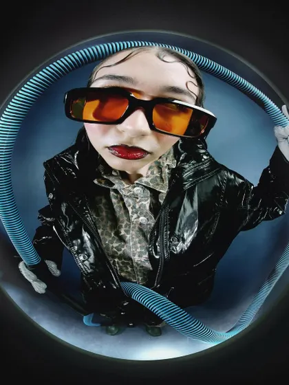 A fisheye portrait with the model in orange sunglasses, framed by a blue corrugated tube. The composition is playful and futuristic, using bold colors and shapes to create a memorable image.