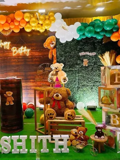 A charming teddy bear-themed birthday party. The setup uses a wood-paneled backdrop, a greenery wall, and an orange, green, and gold balloon arch to create a warm, rustic feel.