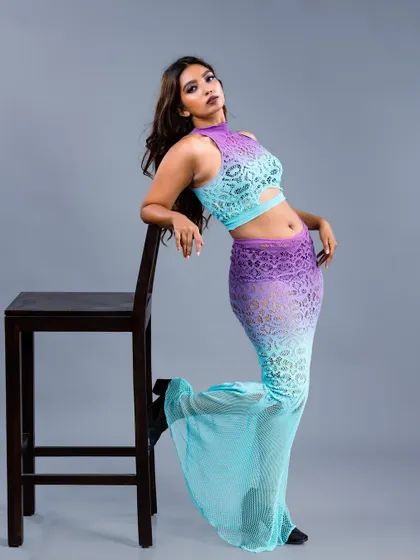 A model in a unique purple and blue ombre lace two-piece outfit, leaning against a stool. The colors and texture make this a standout look for an editorial shoot.