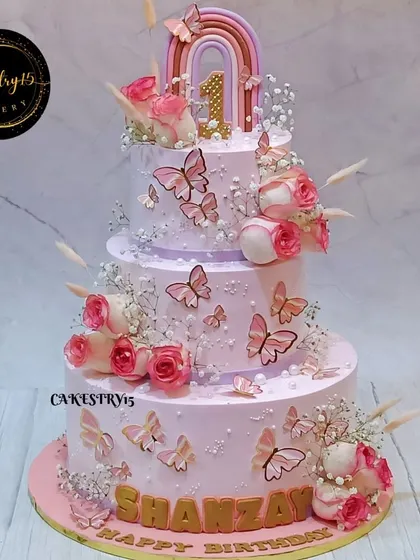 A grand three-tier butterfly-themed cake for a first birthday. This stunning creation is adorned with fluttering butterflies, real pink roses, and a fondant rainbow topper.