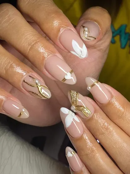 Elegant and sophisticated. These almond nails feature a nude base with delicate white floral art, gold lines, and pearl accents.