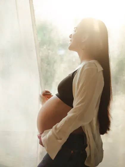 A beautiful backlit portrait of a mother-to-be in a casual outfit. The natural light from the window creates a warm, ethereal glow.