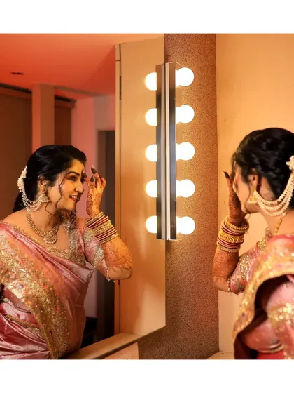 A beautiful shot of the bride getting ready for her engagement, looking at her reflection in a mirror with vanity lights. It's a classic and glamorous pre-ceremony portrait.