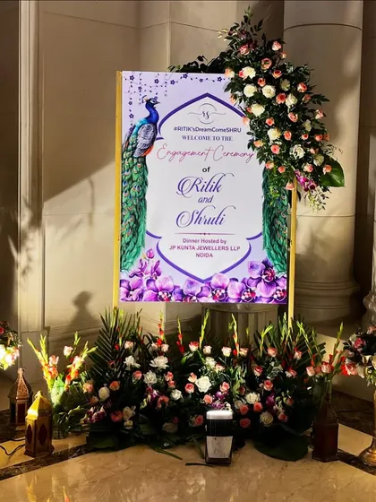 A grand welcome sign for Ritik and Shruti's engagement at The Leela Palace. The design features a beautiful peacock motif and is surrounded by a lavish arrangement of fresh flowers.