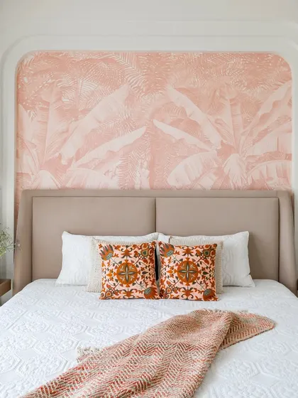A serene and stylish bedroom featuring a custom upholstered bed set against a soft pink, palm-leaf patterned accent wall. The design is completed with elegant pendant lights and carefully chosen textiles to create a tranquil retreat.