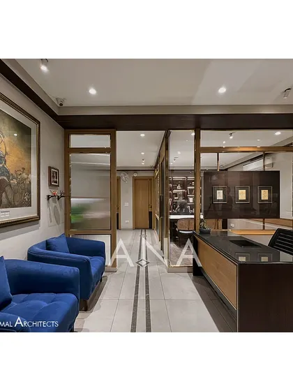 The reception and waiting area of the Jain Cosphere Office. The design uses a palette of beige, wood, and dark green, with blue sofas adding a pop of color. The space is designed to feel open yet private.