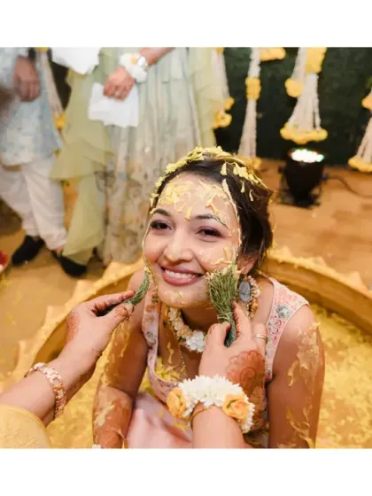 The bride smiles as a leaf is used to gently apply haldi to her face. This image captures the contrast between the messy fun and the tender, loving moments of the ceremony.