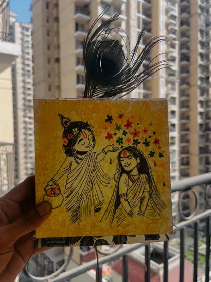 The Radha-Krishna Holi painting held up against a city backdrop, with a peacock feather adding a divine touch.