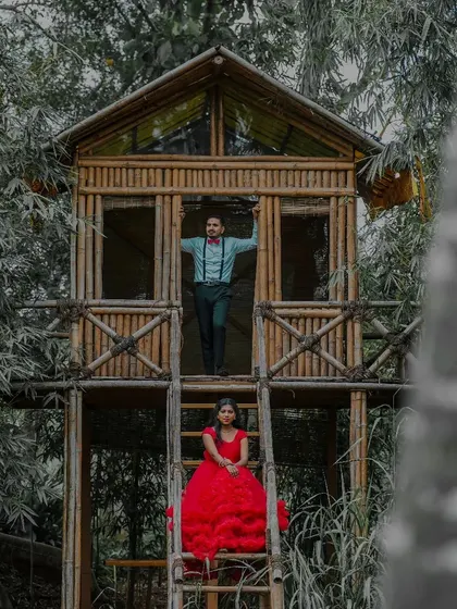 This creative shot features a couple in a treehouse setting. The vibrant red of the ruffled gown stands out dramatically against the rustic, natural tones of the wood and forest.