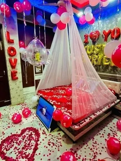 This 'Just Married' room decor is all about celebration. The room is filled with red, pink, and white balloons, with a floor covered in petals, a heart shape on the floor, and a canopy over the decorated bed.