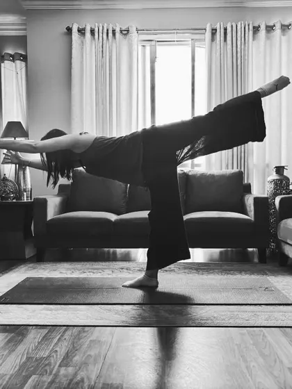 Warrior three in black and white at home, arms back and one leg lifted level with the floor.