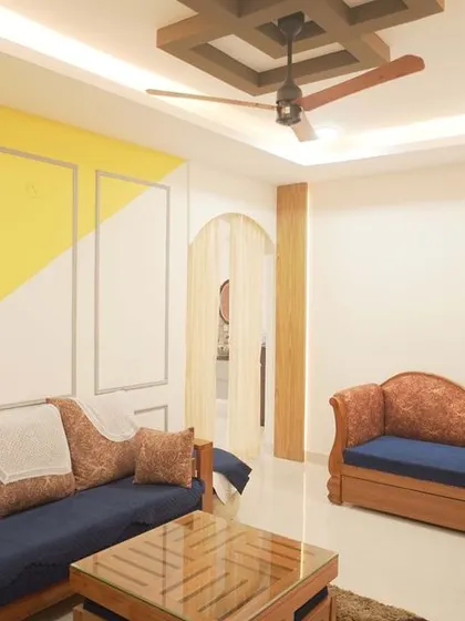A living room that blends traditional and modern styles, with a classic wooden diwan and sofa set against a contemporary accent wall with geometric paintwork. The wooden rafters on the ceiling add a rustic touch.