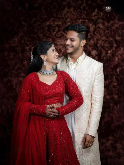 A classic studio-style portrait of a couple dressed for their reception or engagement. The bride in a stunning red lehenga and the groom in an ivory sherwani look at each other with warm smiles.