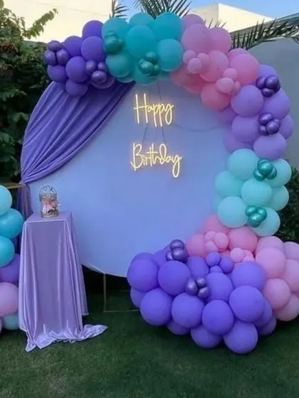An outdoor birthday setup with a circular backdrop and a beautiful balloon arch in shades of purple, pink, and teal, finished with a 'Happy Birthday' neon sign.