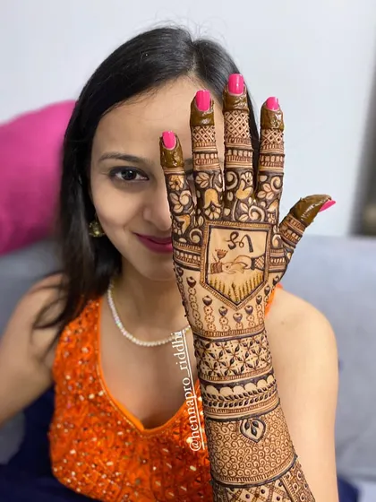 A happy bride peeking through her henna-adorned hand. This back-hand design features a personalized shield with the couple's initials and a "gathbandhan" knot symbol.