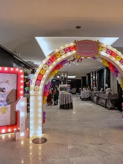 A grand carnival entrance archway for an indoor event. The arch is decorated with colorful balloons and marquee lights, flanked by light-up signs and a friendly elephant prop to welcome guests to the celebration.