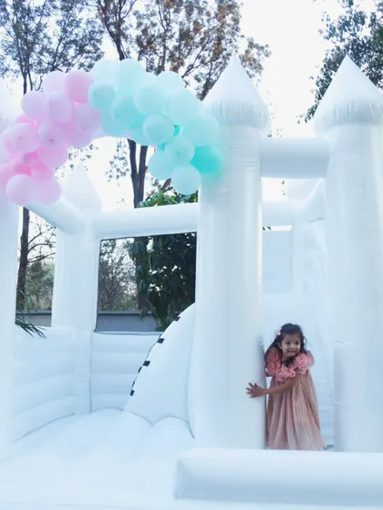 The birthday girl peeks out shyly from the entrance of the beautiful all-white bouncy castle, adorned with a simple pastel balloon arch.