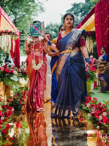 A bride's journey is a cherished family affair. Here, she walks down the golden aisle, accompanied by relatives performing a timeless ritual, all while surrounded by vibrant red floral decor that symbolizes love and celebration.