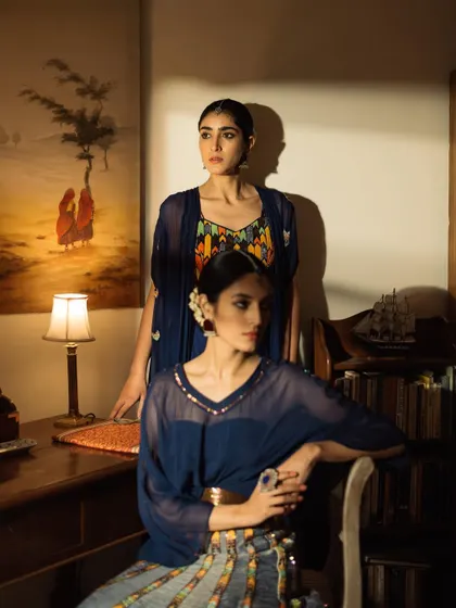 A moody, atmospheric shot from a lookbook, featuring two models in a dimly lit, studious room. The lighting is cinematic, highlighting the deep blue tones of the outfits.