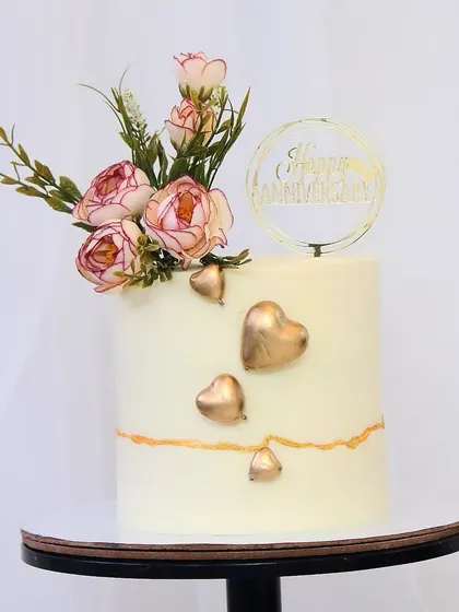 A simple yet beautiful anniversary cake. The clean white buttercream is accented with three-dimensional golden hearts and a small, elegant floral arrangement on top.