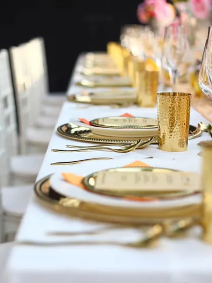 A long dining table set with gleaming gold-plated cutlery and glasses against crisp white linens. This modern and luxurious setup is perfect for an elegant wedding reception or corporate gala.