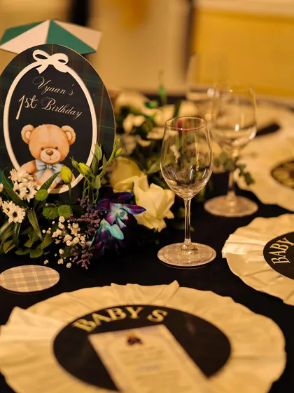 A detailed view of the personalized table sign for "Vyaan's 1st Birthday," featuring the adorable teddy bear motif. The surrounding florals and elegant glassware showcase our commitment to a polished and complete look.
