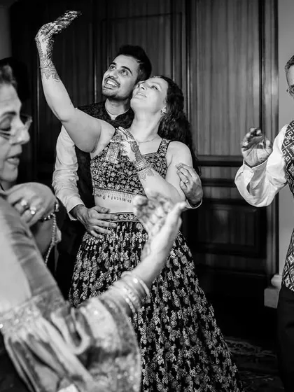 A multi-generational dance moment. The couple takes a selfie while their parents dance around them, a beautiful blend of modern and traditional.
