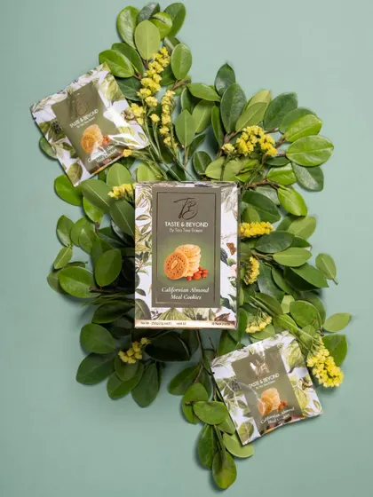 A creative flat lay of Taste & Beyond cookie packaging arranged on a bed of fresh green leaves. This styling emphasizes the natural ingredients and freshness of the product.