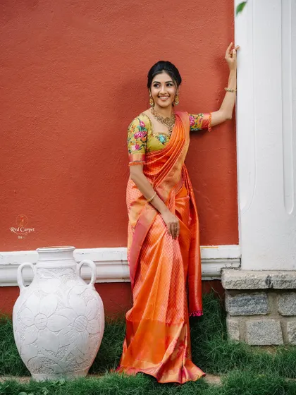 The vibrant orange of the saree against the rustic red wall creates a beautiful, warm color palette.