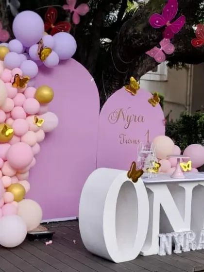 An outdoor butterfly-themed birthday setup, using a large tree as a natural part of the decor. The arrangement includes pastel balloon arches and large "ONE" lettering.
