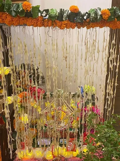 A small mandap created for a home pooja. The frame is wrapped in marigold garlands, and strings of rajnigandha (tuberose) hang in the background for a traditional look and fragrance.