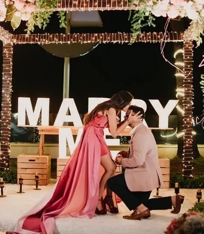 A tender moment after she says yes, framed by the romantic decor and the 'MARRY ME' lights.