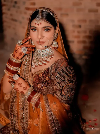 A striking portrait focusing on the bride's intense gaze and henna-adorned hands. The warm tones of the photo enhance the richness of her brown and gold lehenga.
