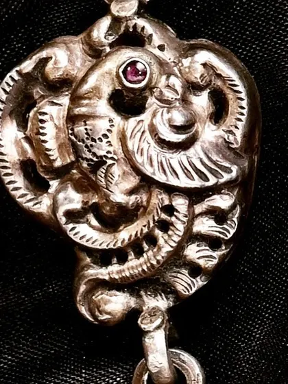 A close-up of the handcrafted silver peacock pendant, showing the fine details of the carving against a dark background.