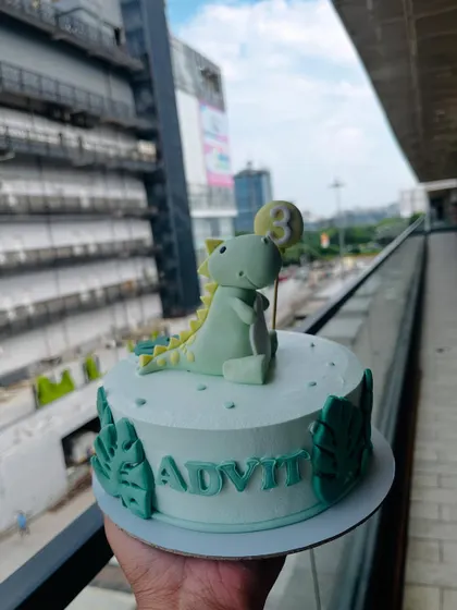 A simple and sweet dinosaur cake for a third birthday. This design features a cute, green fondant dinosaur holding a balloon, surrounded by monstera leaves.