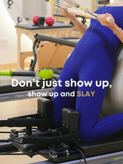 This is our philosophy: show up to slay. A client demonstrates a kneeling lat pulldown on the reformer, using a stability ball to engage her core while strengthening her back muscles.
