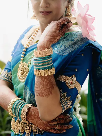 A detailed shot of the bride's arm, showcasing her intricate henna, temple jewelry-inspired armlet (vanki), and traditional bangles against her vibrant blue and green saree.