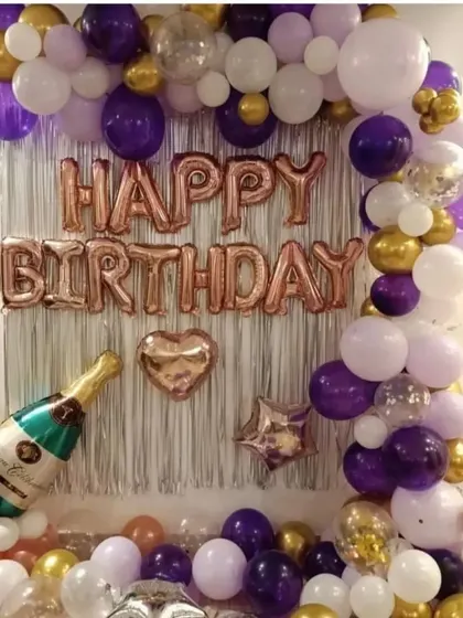 A simple birthday decoration with a purple, gold, and white balloon garland against a shimmer curtain.
