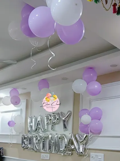 A simple birthday setup with silver 'Happy Birthday' foil letters and bunches of purple and white balloons on the ceiling.