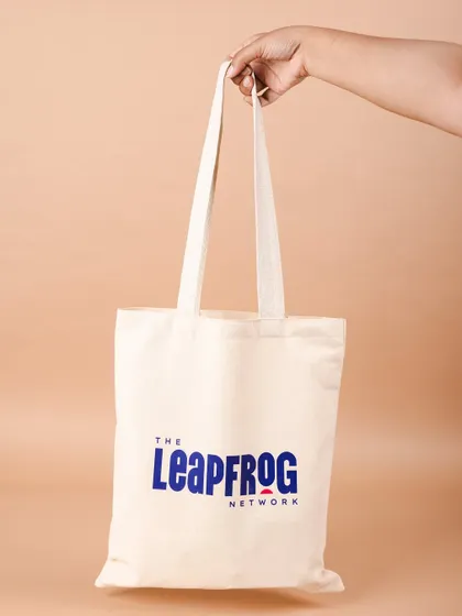 A hand holding up the branded tote bag for The Leapfrog Network. We love how clean and impactful the logo looks on the canvas.