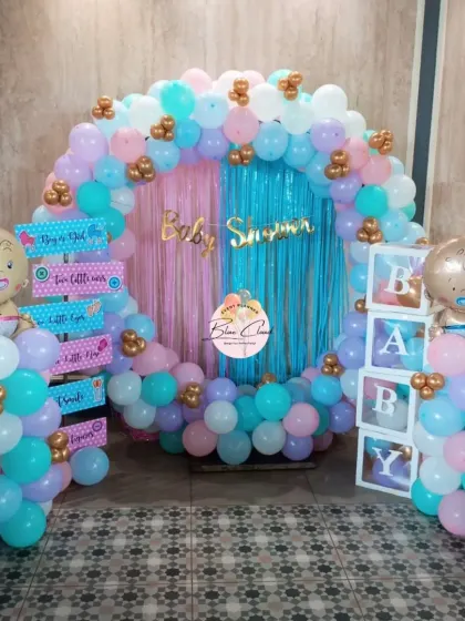 A beautiful gender reveal party setup with a full balloon ring in pastel pink, blue, and purple. The decor includes 'BABY' blocks, themed foil balloons, and a shimmer curtain backdrop.