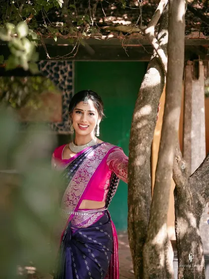 A smiling bride in a stunning pink and blue saree, posing naturally by a tree.