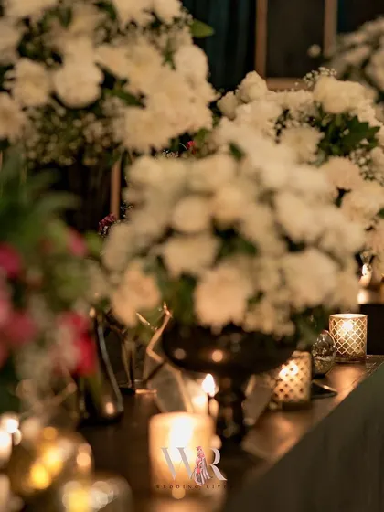A close-up of white floral arrangements in dark vases, with the soft, warm light of candles creating a moody and intimate atmosphere.