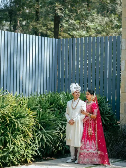 A full-length portrait of the couple in their wedding attire. The bride's bright pink lehenga stands out against the lush greenery and modern architecture.