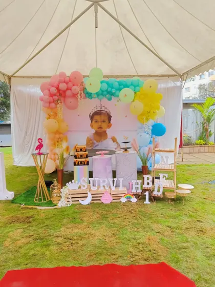 An outdoor party setup under a canopy for Survi's first birthday. The decoration features a pastel rainbow balloon arch and a large photo of the birthday girl.