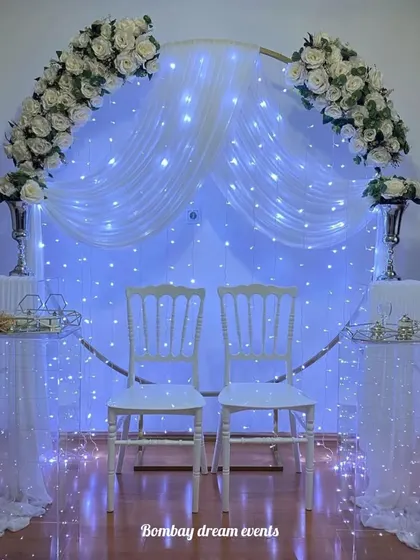An elegant and simple anniversary backdrop. This setup features a circular frame draped with white fabric, fairy lights, and white floral arrangements, creating a perfect photo opportunity for the happy couple.