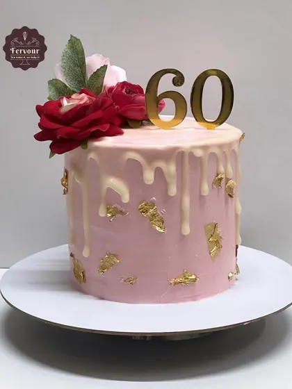 A drip cake designed for a 60th birthday celebration. The pink frosting is adorned with a white chocolate drip, gold leaf, and fresh red roses for a classic and beautiful look.