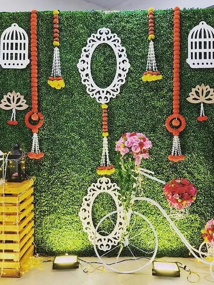 A creative photo backdrop for a Mehendi. The green turf wall is decorated with white frames, birdcages, and a decorative bicycle, creating a charming and rustic look.