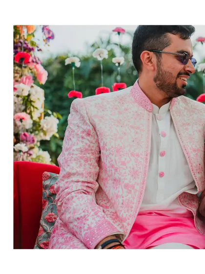 The groom looking dapper in his pink and white sherwani against a floral backdrop. A candid smile captured during the vibrant Mehendi festivities.