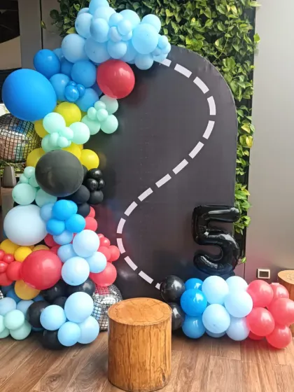 A racetrack-themed decoration for a 5th birthday. The setup includes a black arched backdrop with a road graphic and a colorful balloon garland.